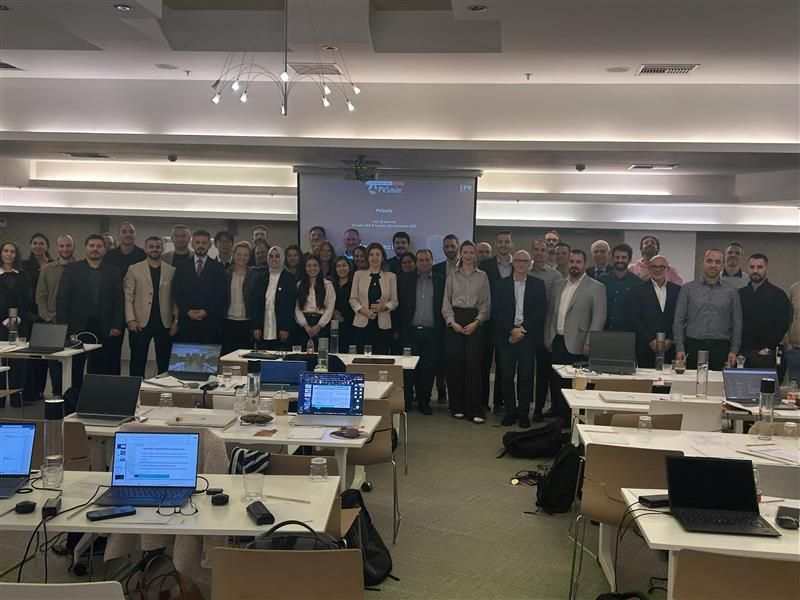 Large group of PVSmile project partners posing for a photo in a conference room, standing in front of a projector screen, with tables in the foreground covered with open laptops and meeting materials.